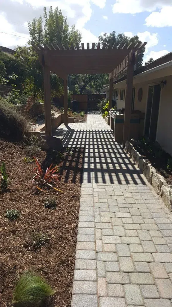 A brick pathway leads to a wooden pergola, casting shadows on the ground.