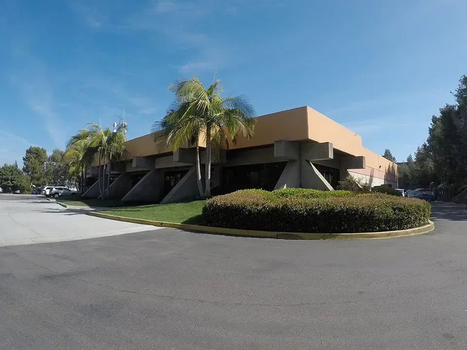 Tan commercial building with angled supports, palm trees, and a circular hedge on a sunny day.