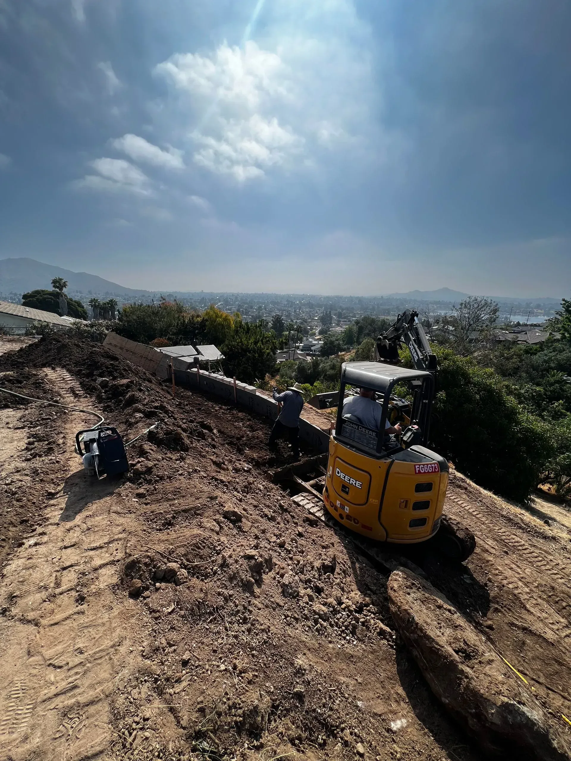 Yellow excavator digging a trench along a hillside with a cityscape in the background under a blue sky.