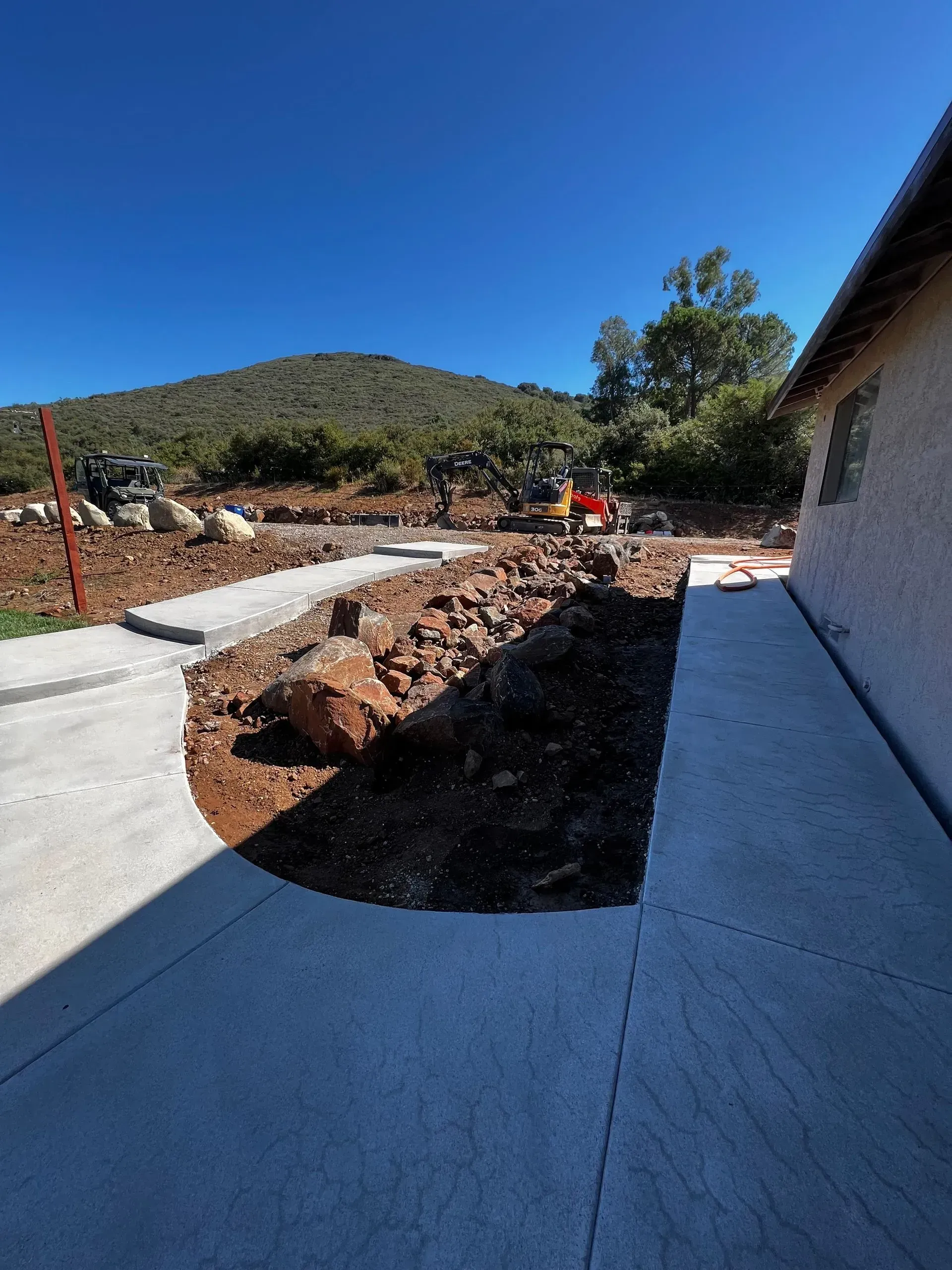 Concrete walkway next to a building and unpaved area with an excavator, mountain in the background.