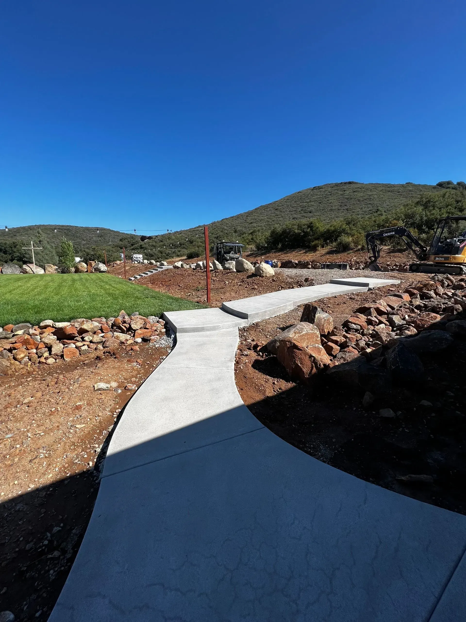 A winding concrete pathway through a landscape with rocks, dirt, grass, and a hill under a blue sky.