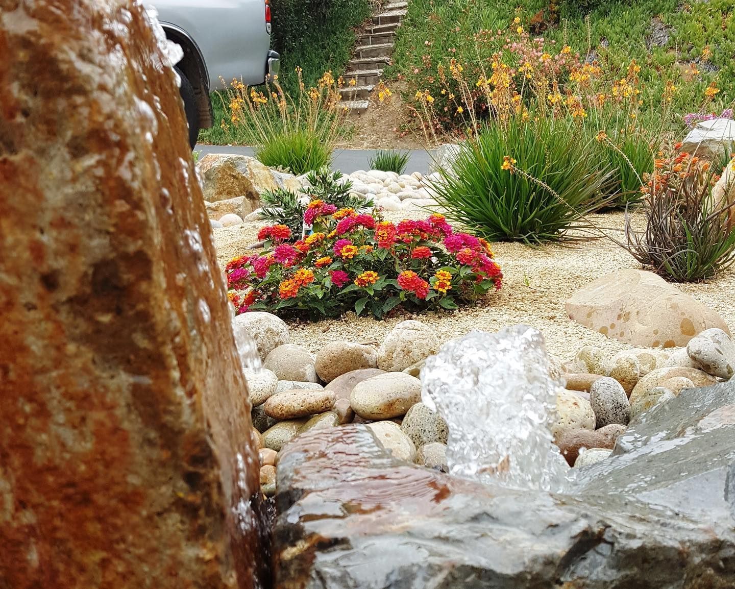Water fountain in a rock garden with colorful flowers and tan stones.