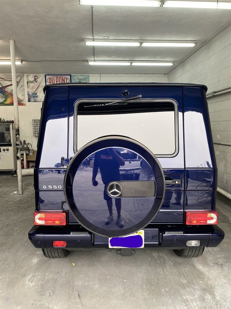 Blue Mercedes-Benz G-Class SUV, rear view, spare tire, person reflected in tire, inside a garage.