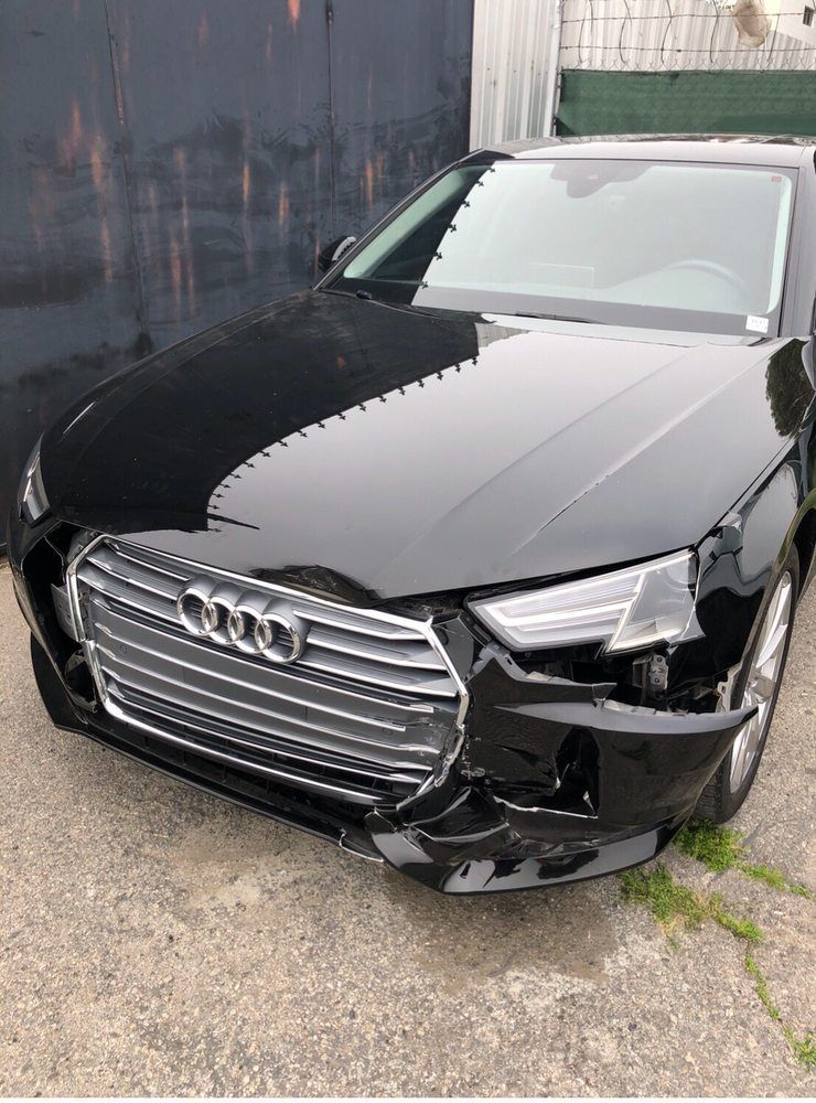 Black Audi car with front-end damage; parked next to a wall.