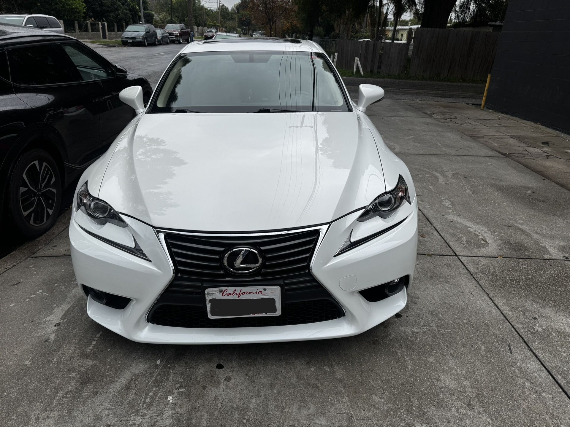 White Lexus sedan parked on a concrete surface, facing forward. Dark grille, headlights, and California license plate visible.