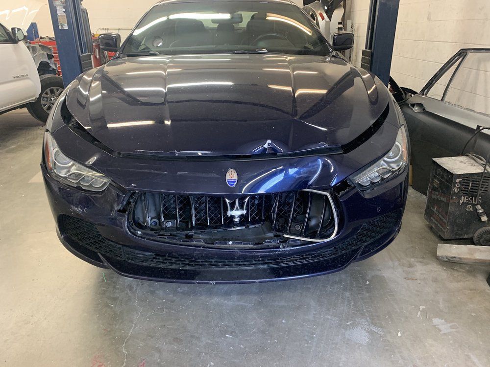Blue Maserati sedan in a garage, front-end damage to the bumper and hood, grill visible.