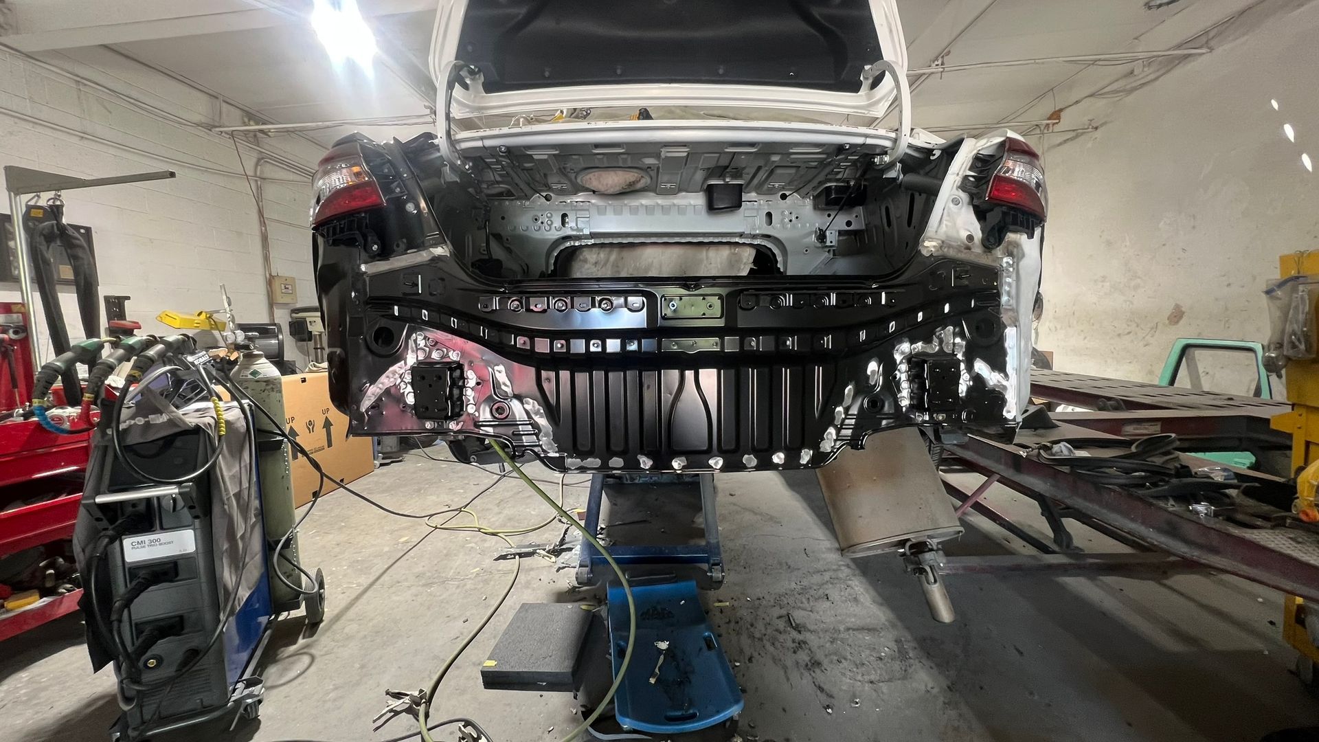 Rear of a car being repaired in a garage, body on a lift. Black and white metal parts with exposed wiring.