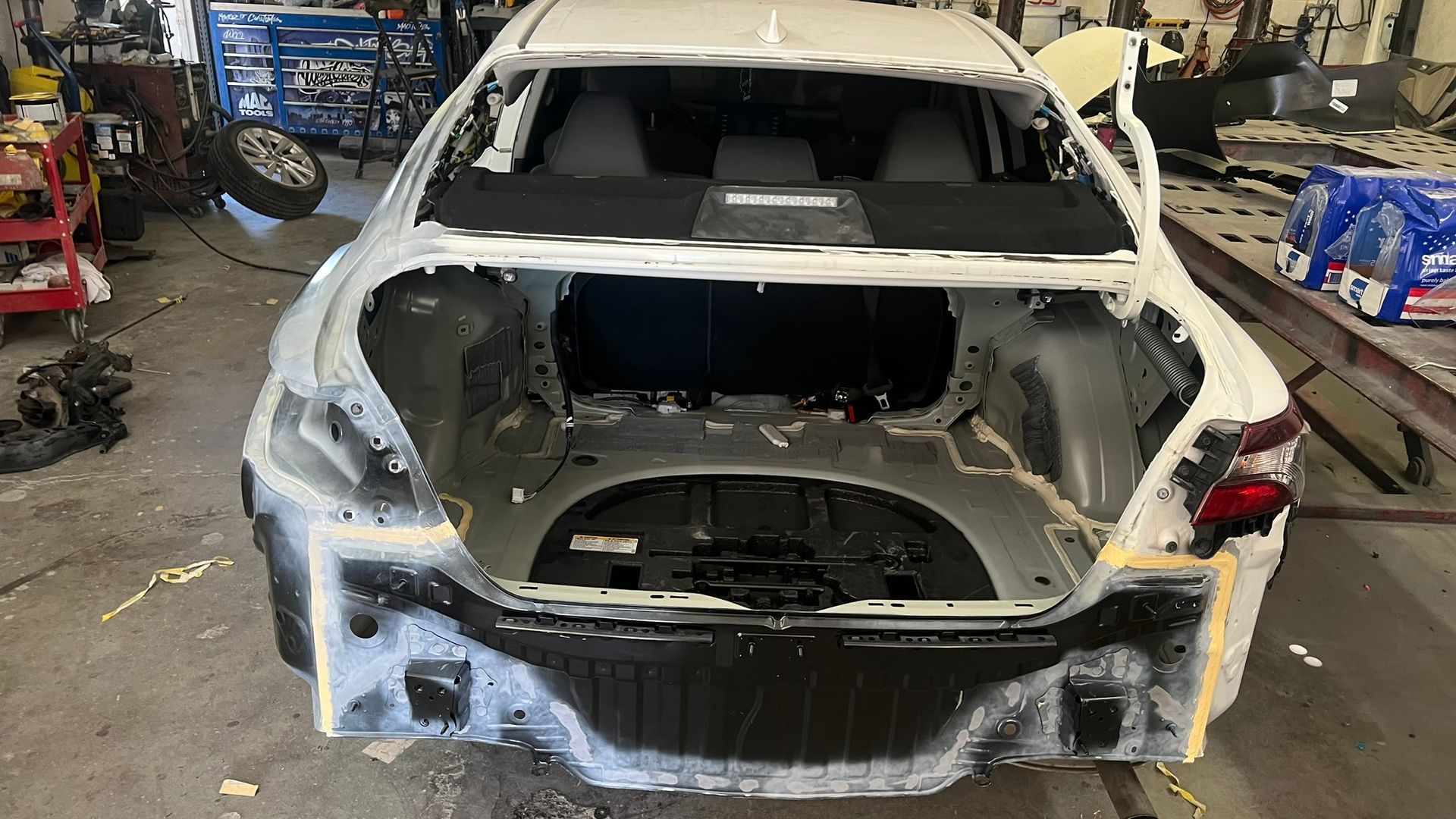 Rear view of a white car undergoing body repair in a garage, revealing the internal structure and trunk.