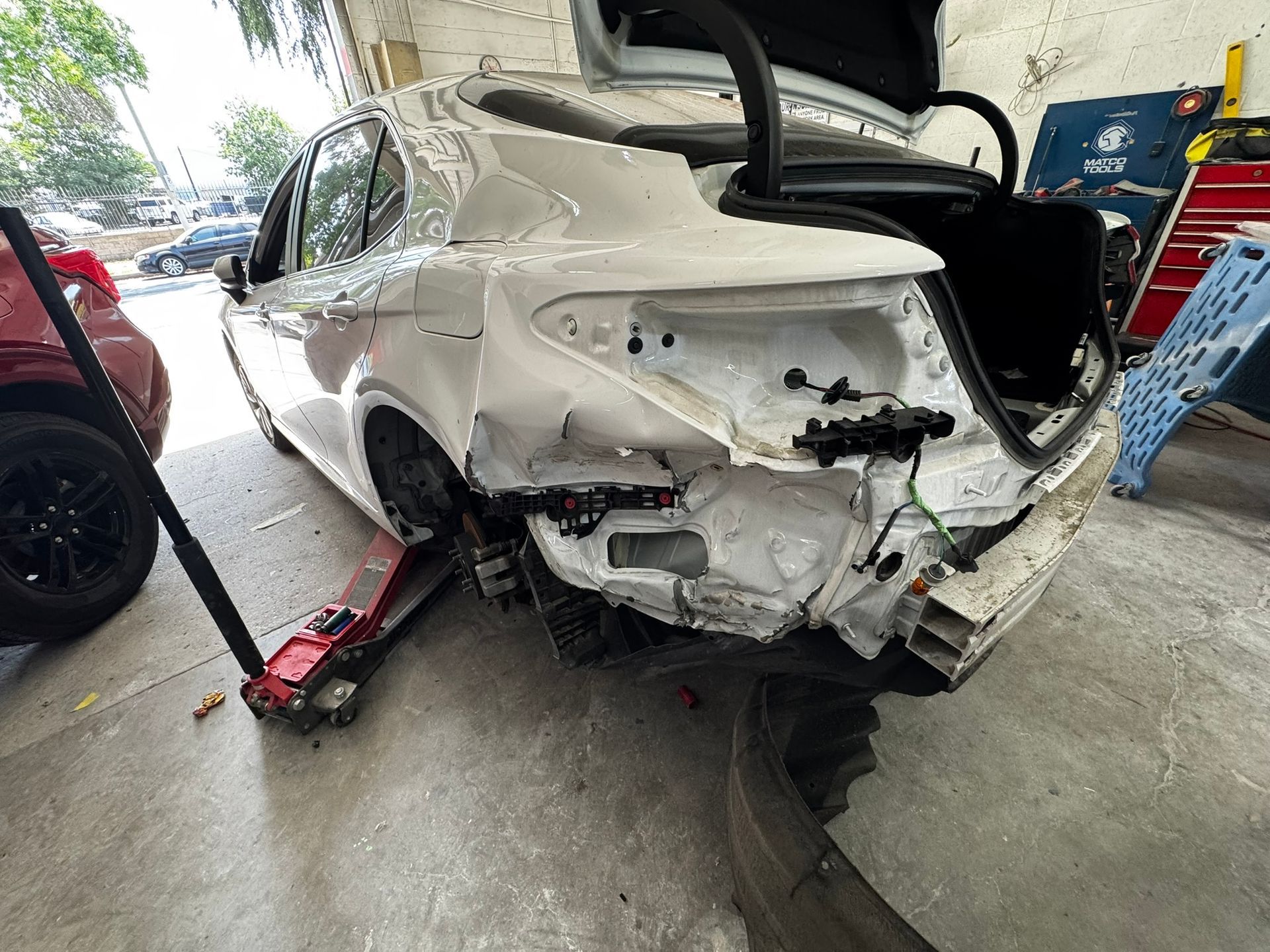 Damaged white sedan in a repair shop, rear end heavily damaged, trunk open, red jack underneath.