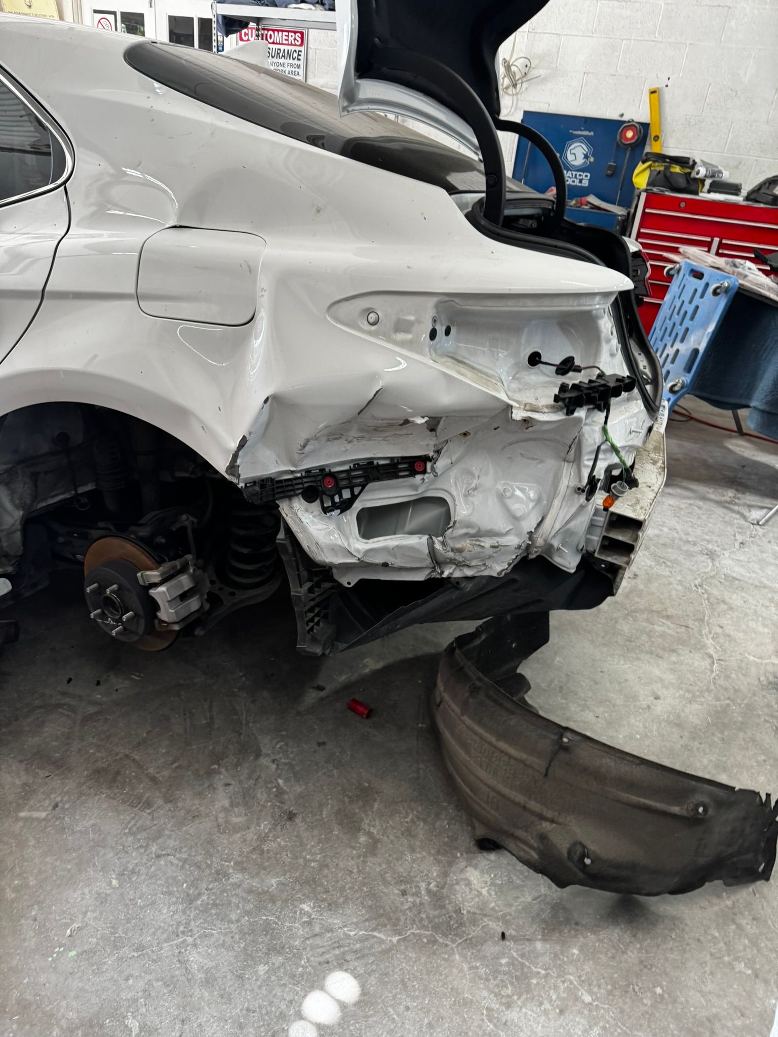 Damaged white car rear end in a repair shop, showing exposed internal components.