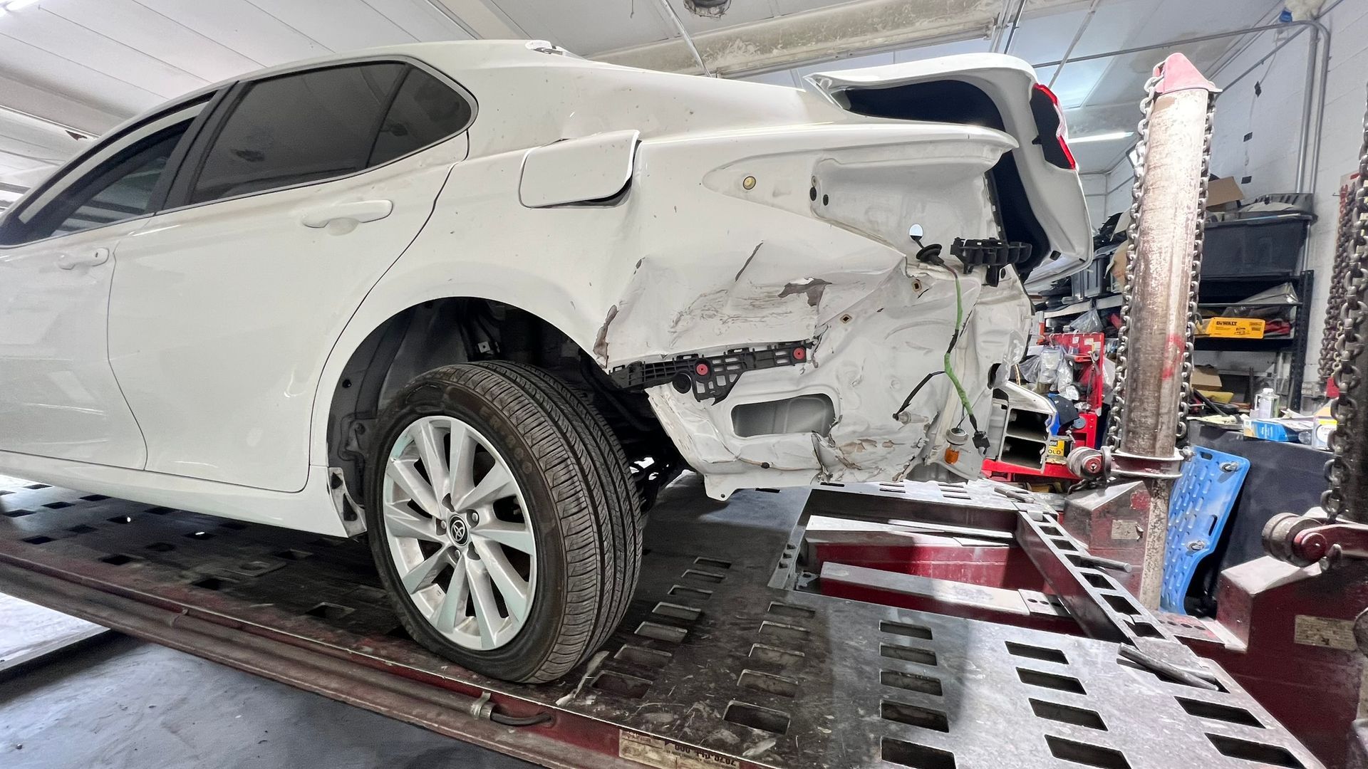 White car with extensive rear-end damage, likely at an auto body shop.