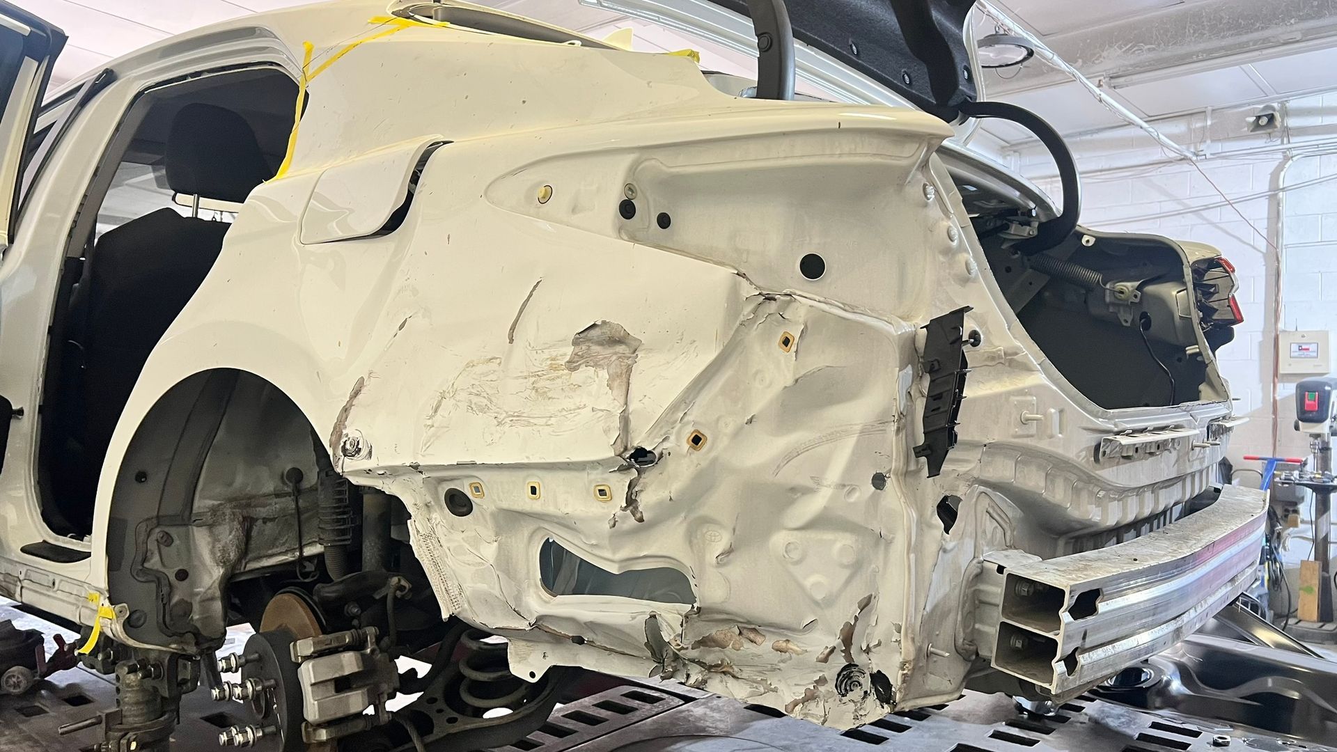 Damaged white car body in auto repair shop, rear end crumpled, trunk open.