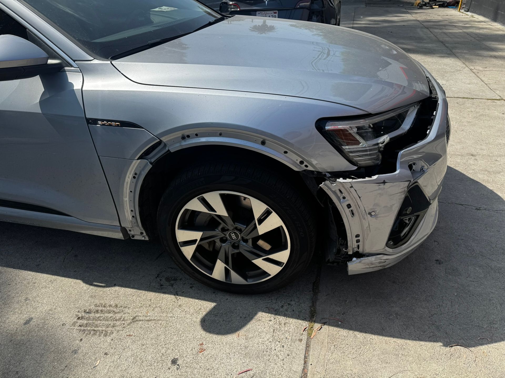 Damaged silver Audi SUV front-end, missing bumper and fender. Tire visible on a paved surface.