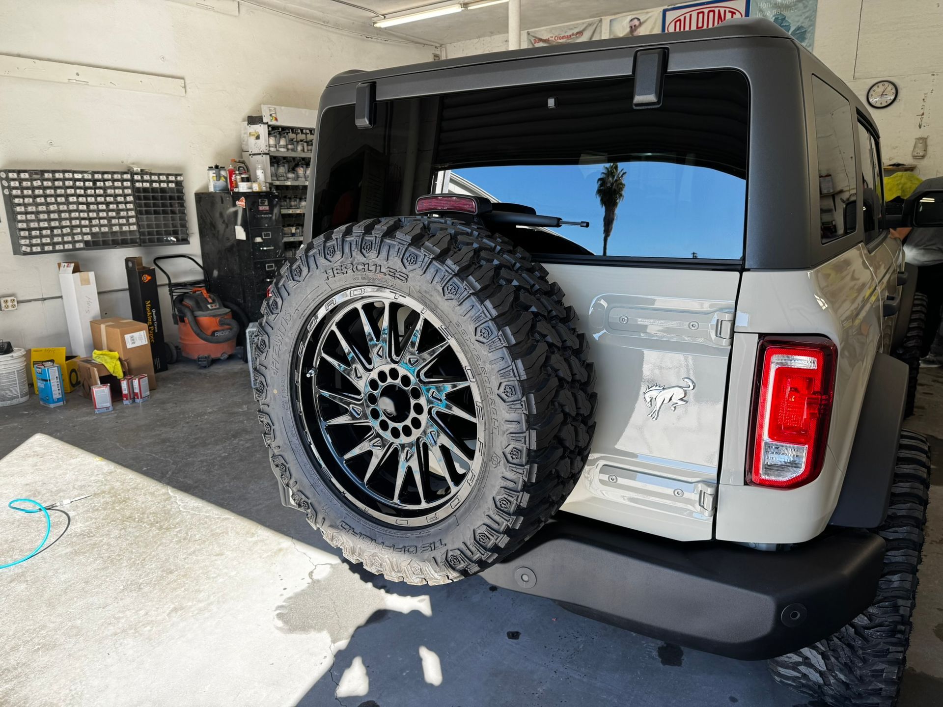 Light tan Ford Bronco with a spare tire on a custom black wheel, in a garage.