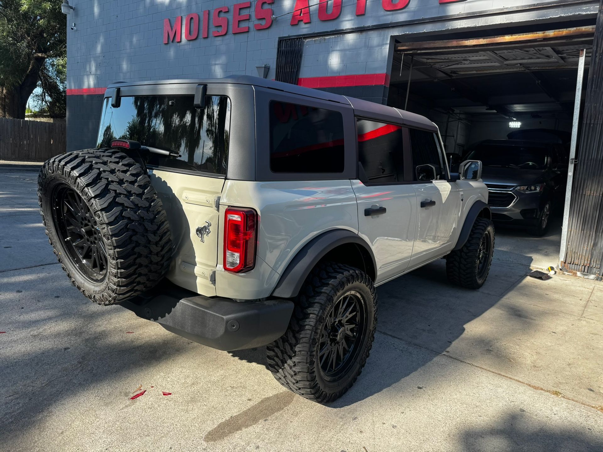 A light tan Ford Bronco with black tires parked at an auto repair shop with the business name 