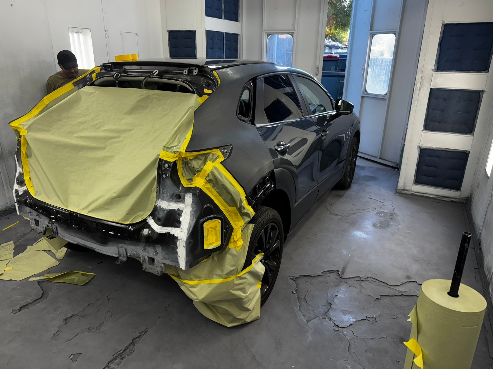 Car in a paint booth, rear end masked with yellow tape and paper for painting. A person is partially visible.