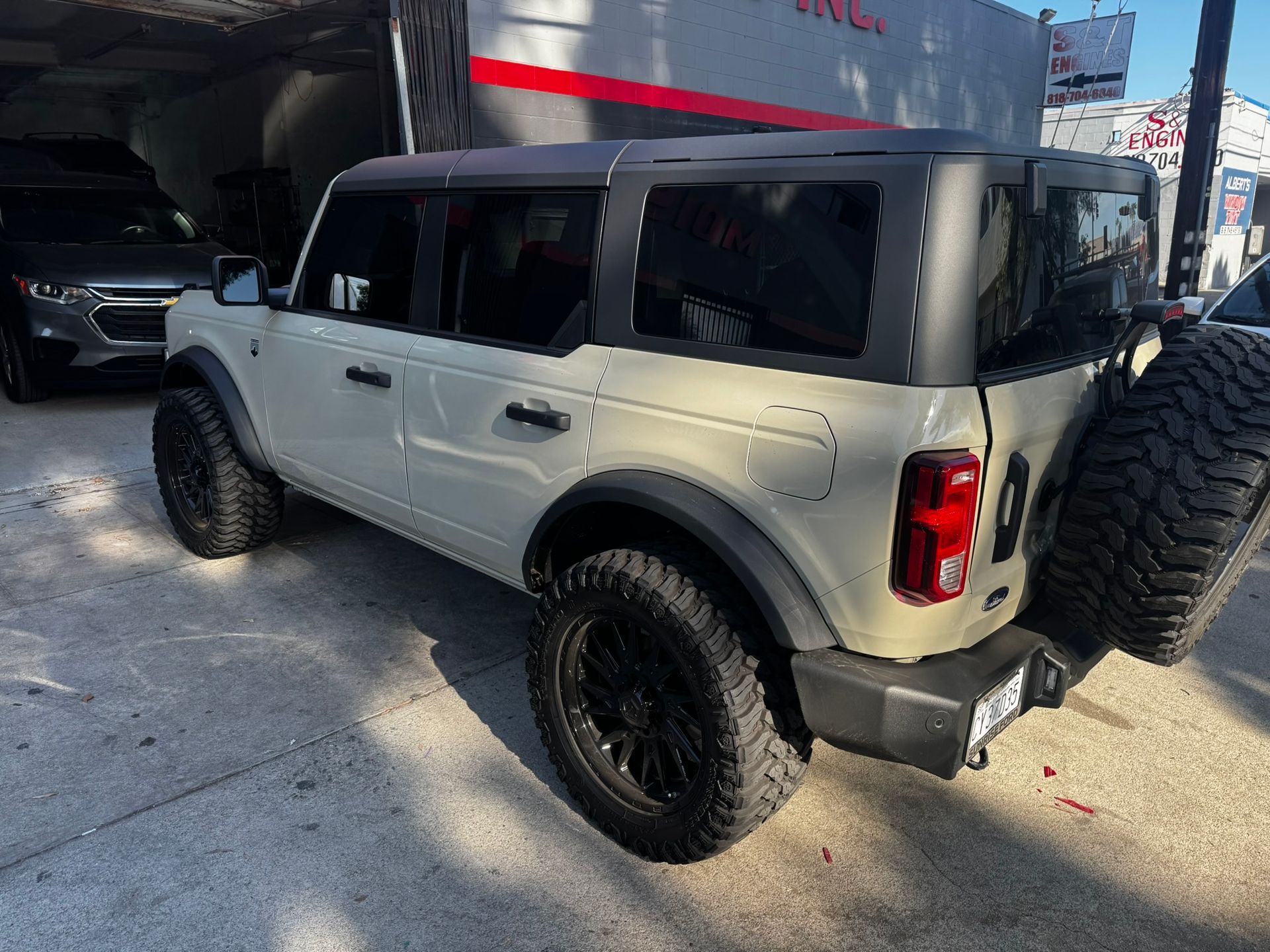Off-white Ford Bronco SUV with black wheels and spare tire parked outside with other vehicles.