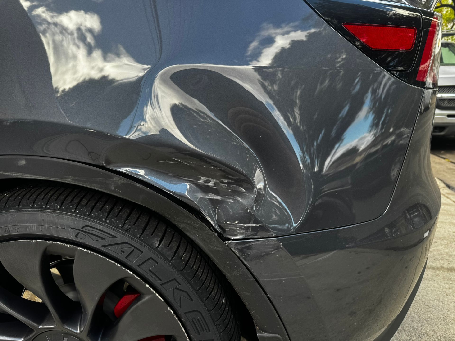 Damaged gray Tesla rear quarter panel with significant dent and scratches. Black wheel and red brake caliper visible.