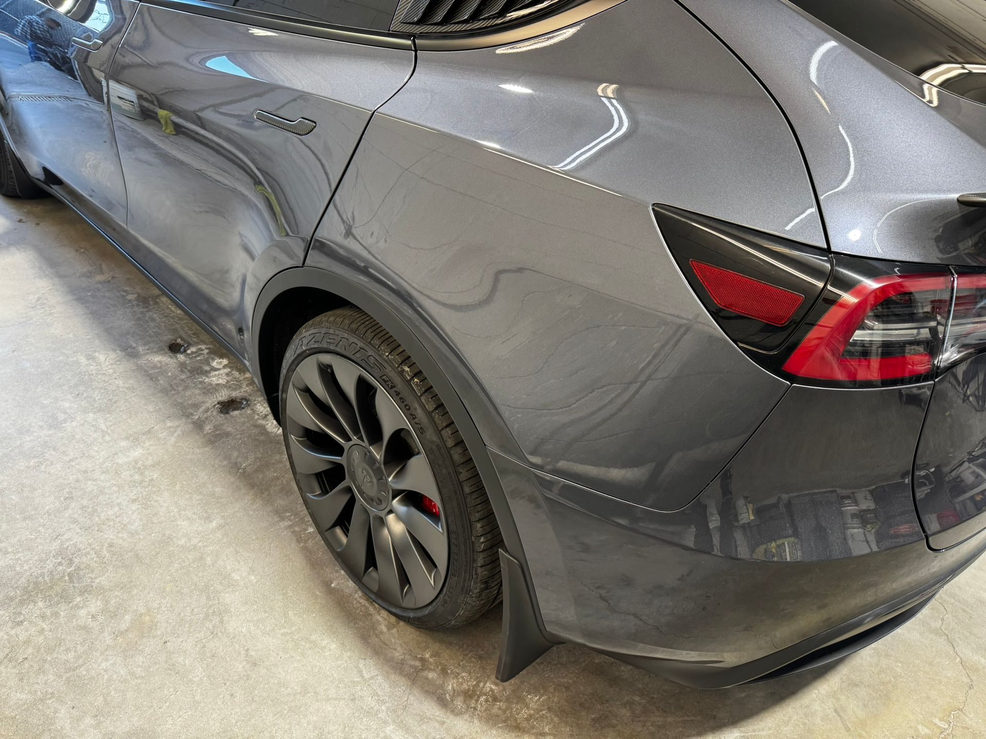 Gray Tesla vehicle with black wheels and taillight, parked indoors on a concrete floor.