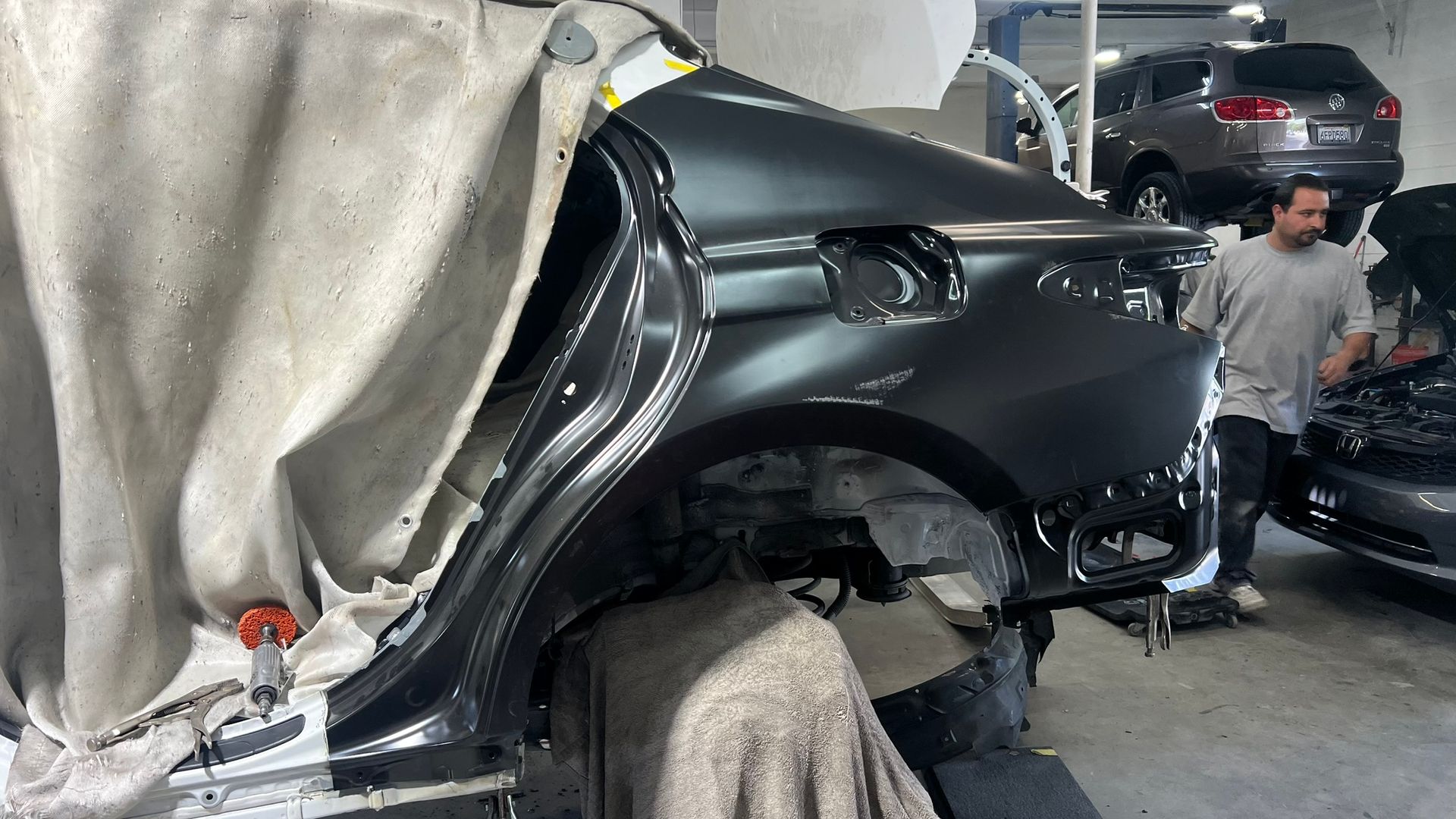 Car being repaired in a body shop; a mechanic stands nearby. The car is dark gray and partially covered.