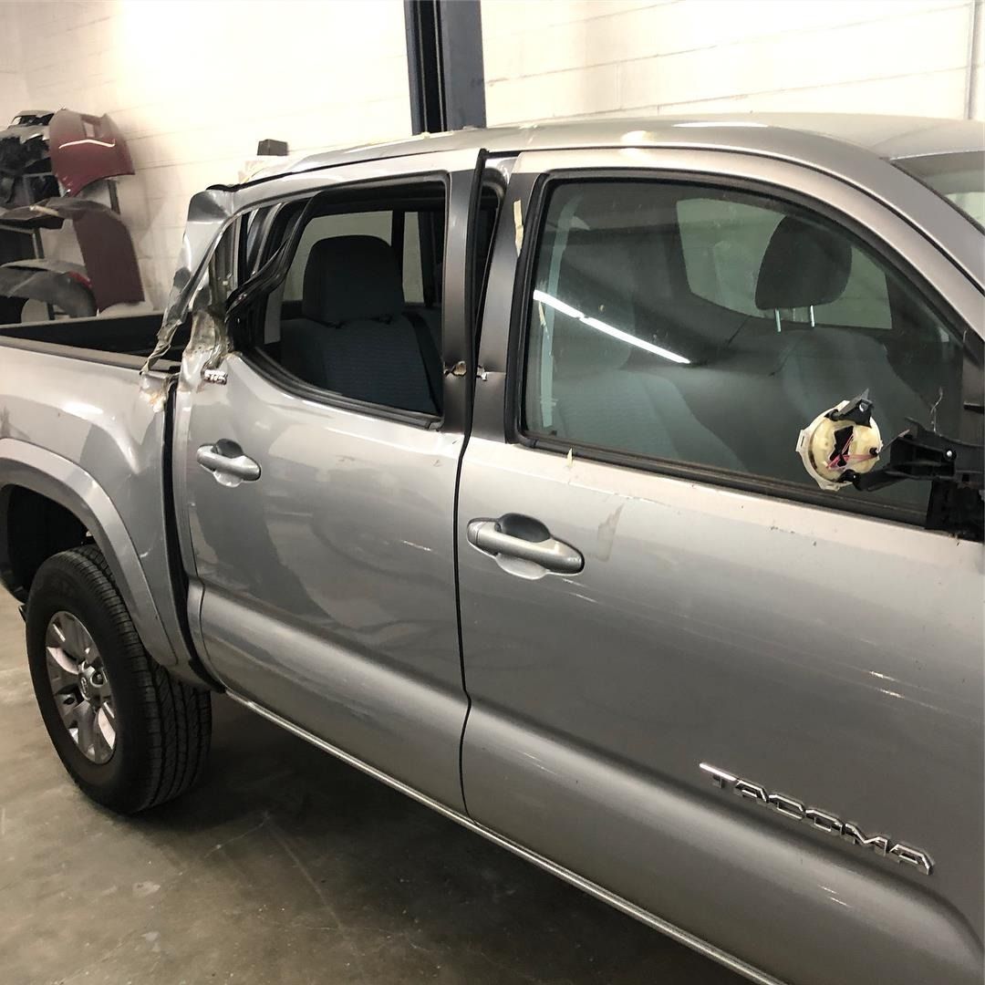 Silver Toyota Tacoma pickup truck in a shop with damaged door and window.