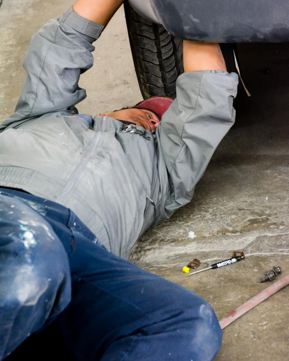 Mechanic in gray jumpsuit working under a vehicle, lying on the floor. Tools nearby.