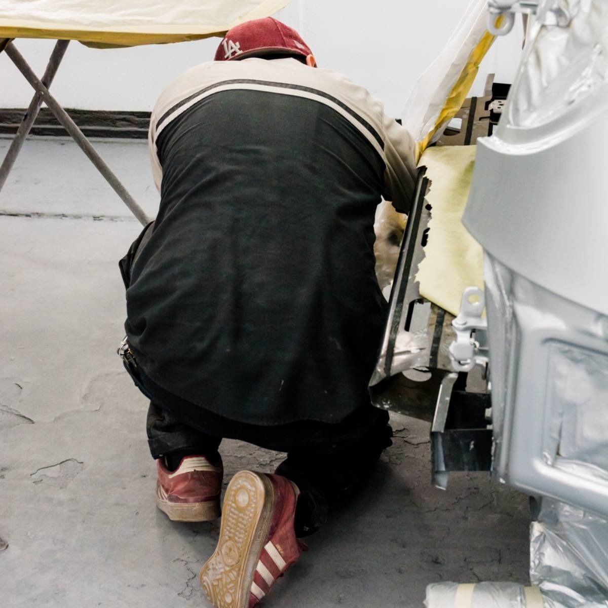 Mechanic kneeling, working on a car part, wearing a cap and black work clothes.