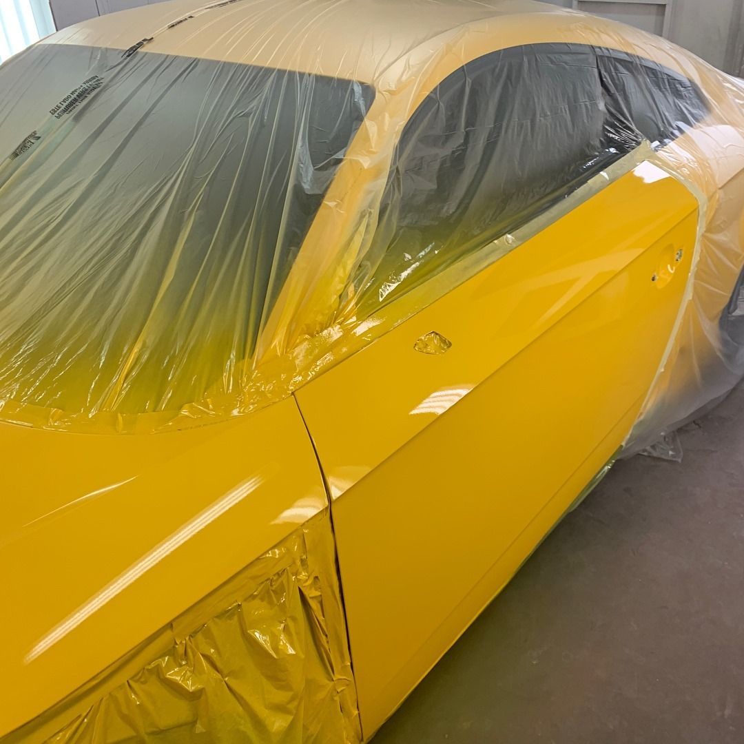 Yellow car being repainted, covered in plastic in a paint booth.
