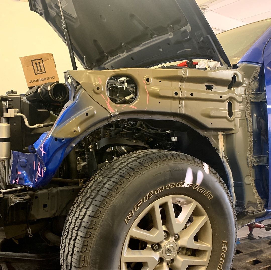Car frame with the hood up, front wheel visible, blue fender, repair shop setting.