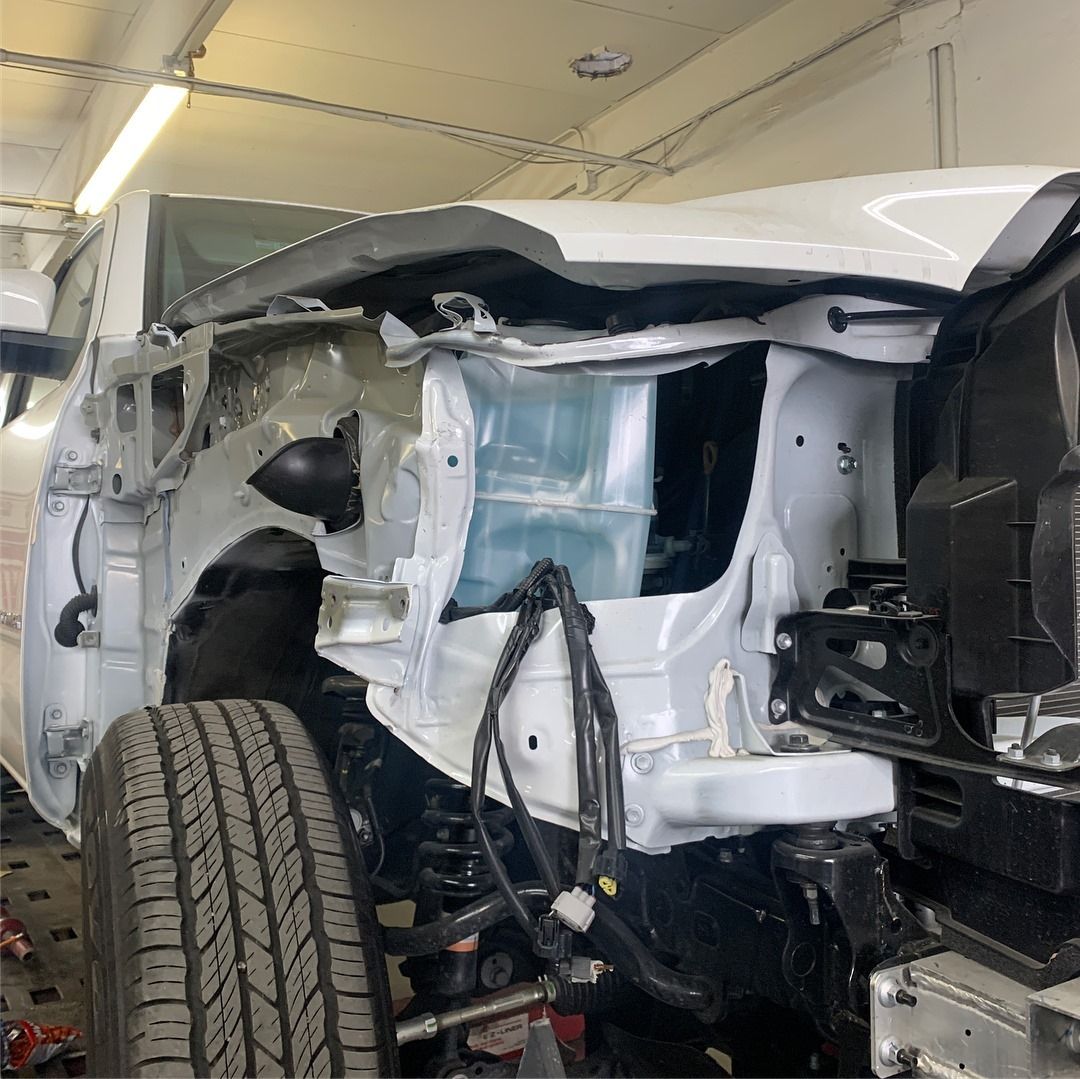 White car front end with damaged bodywork in a repair shop. Tire visible.