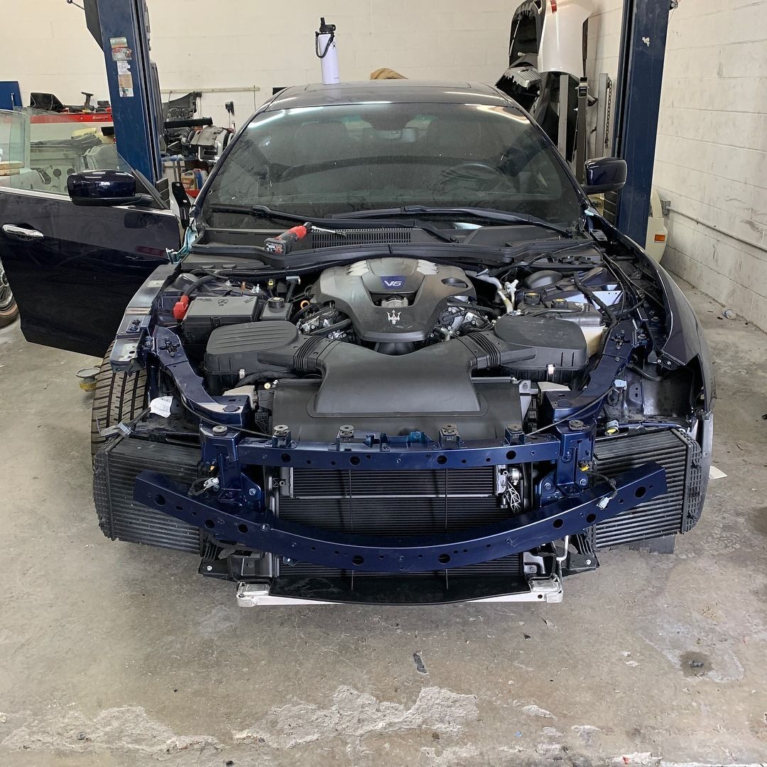 Blue Maserati with open hood in a repair shop, front bumper removed.