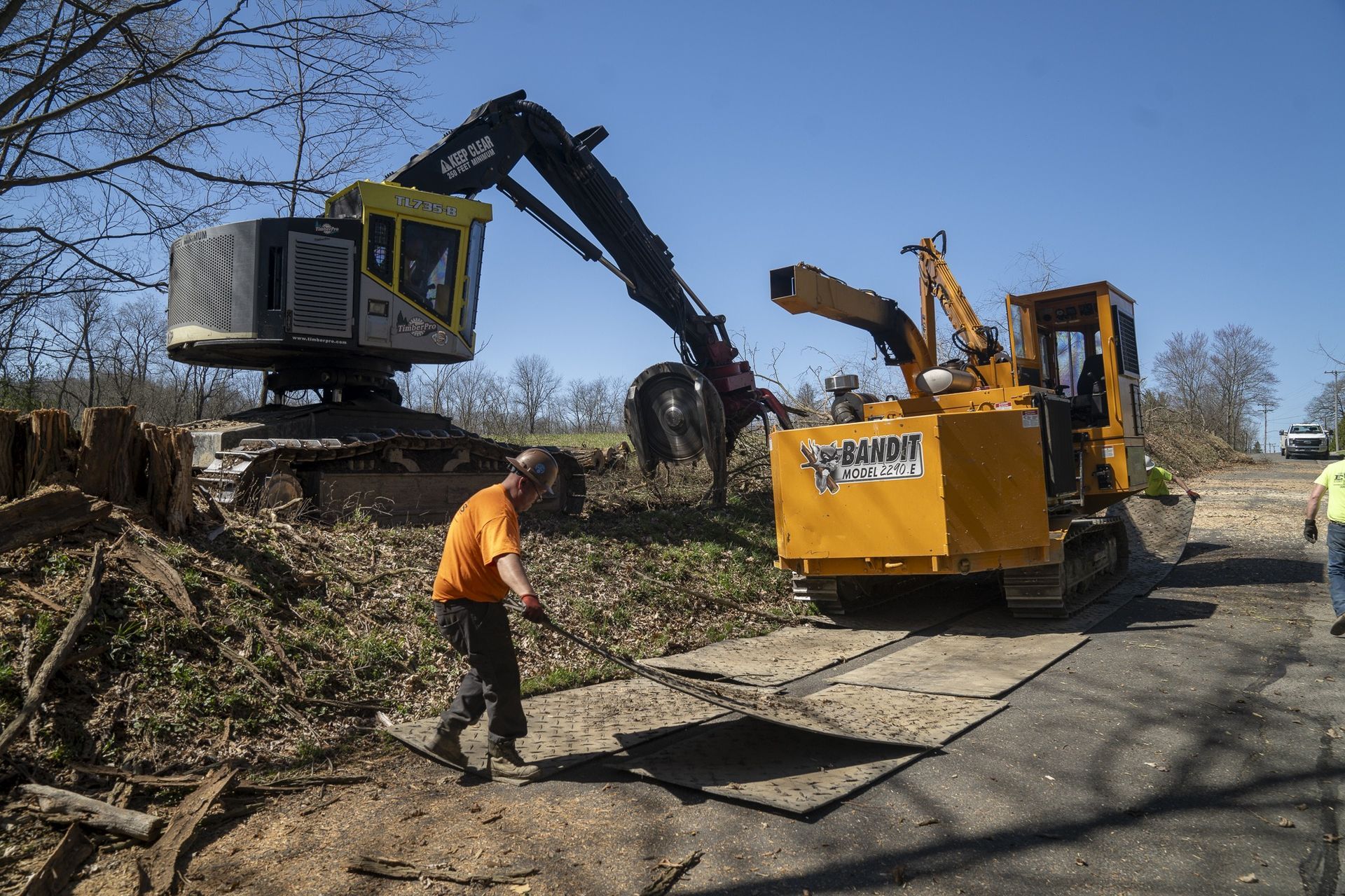 Heavy machinery chipping wood debris in an outdoor setting; a worker in an orange shirt helps.