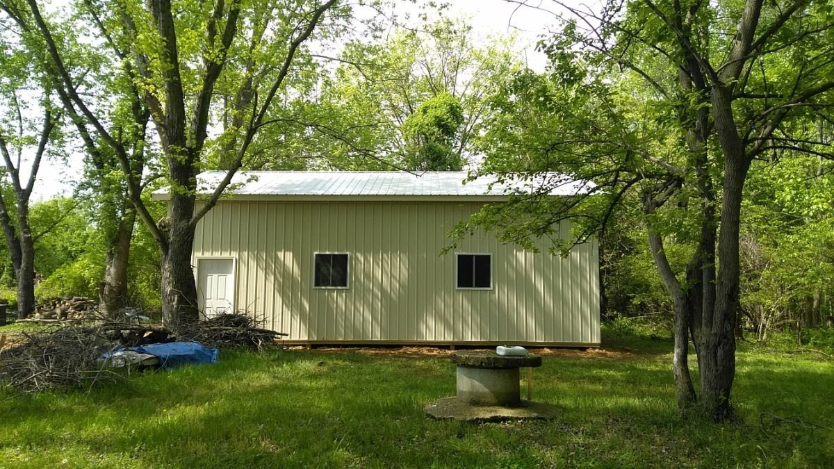 Small beige shed in a wooded yard, with trees and grass surrounding it.