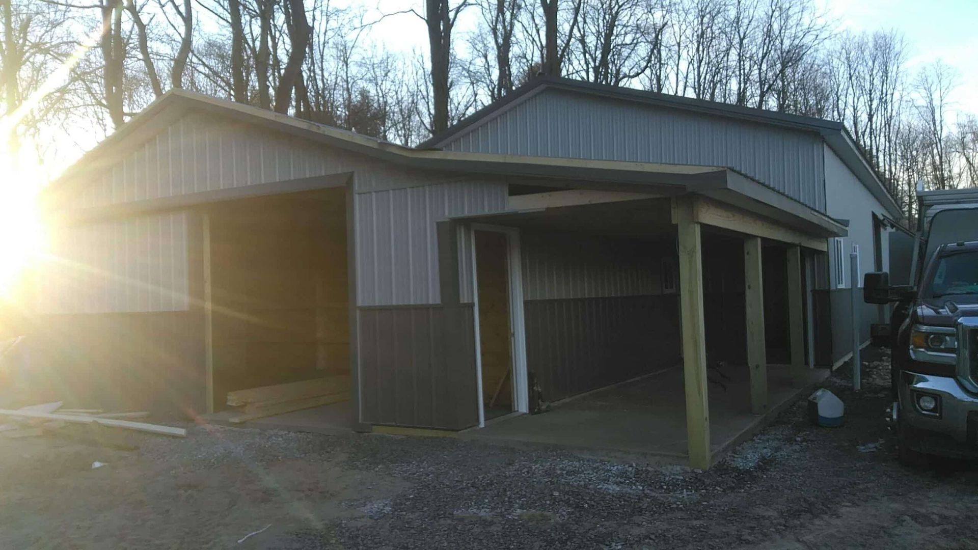 Two-bay metal garage with open doors at sunset, parked truck on the right, trees in the background.