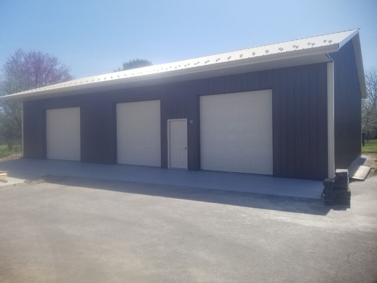 Large dark metal garage with three white roll-up doors on a paved lot under a clear sky