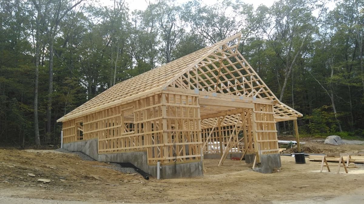Wood-framed house under construction with exposed roof trusses in a wooded area