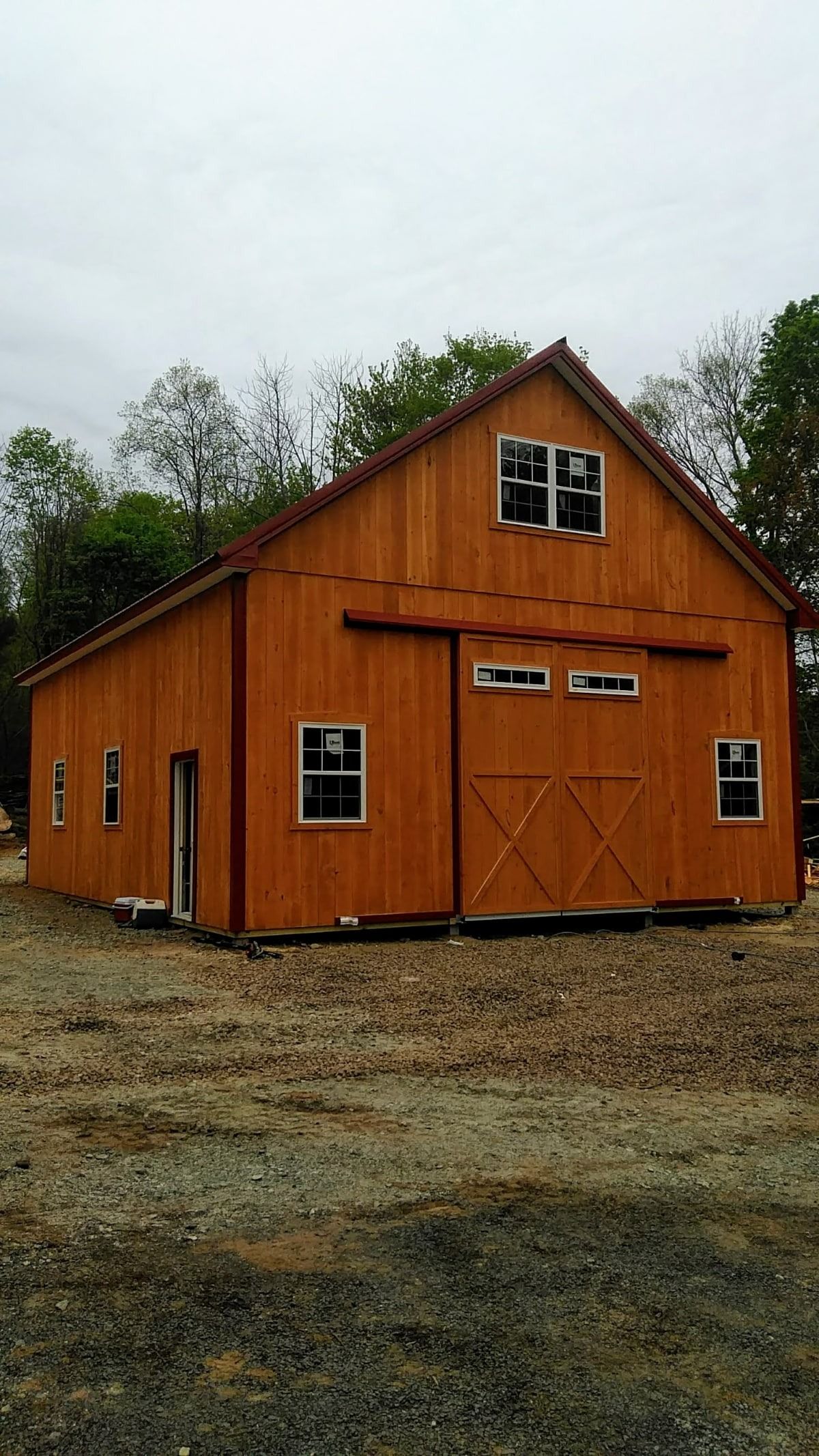 Rustic orange wooden barn with double doors and small windows on a gravel lot, surrounded by trees.