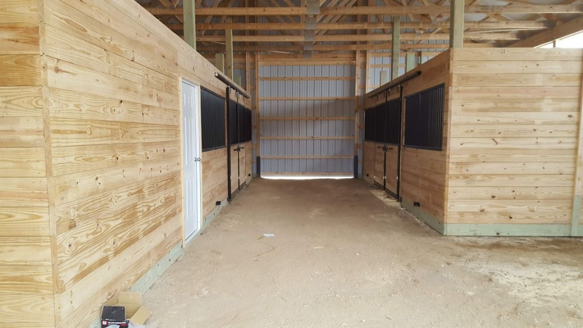 Empty barn aisle with wooden stalls and dirt floor, viewed toward a metal back door.