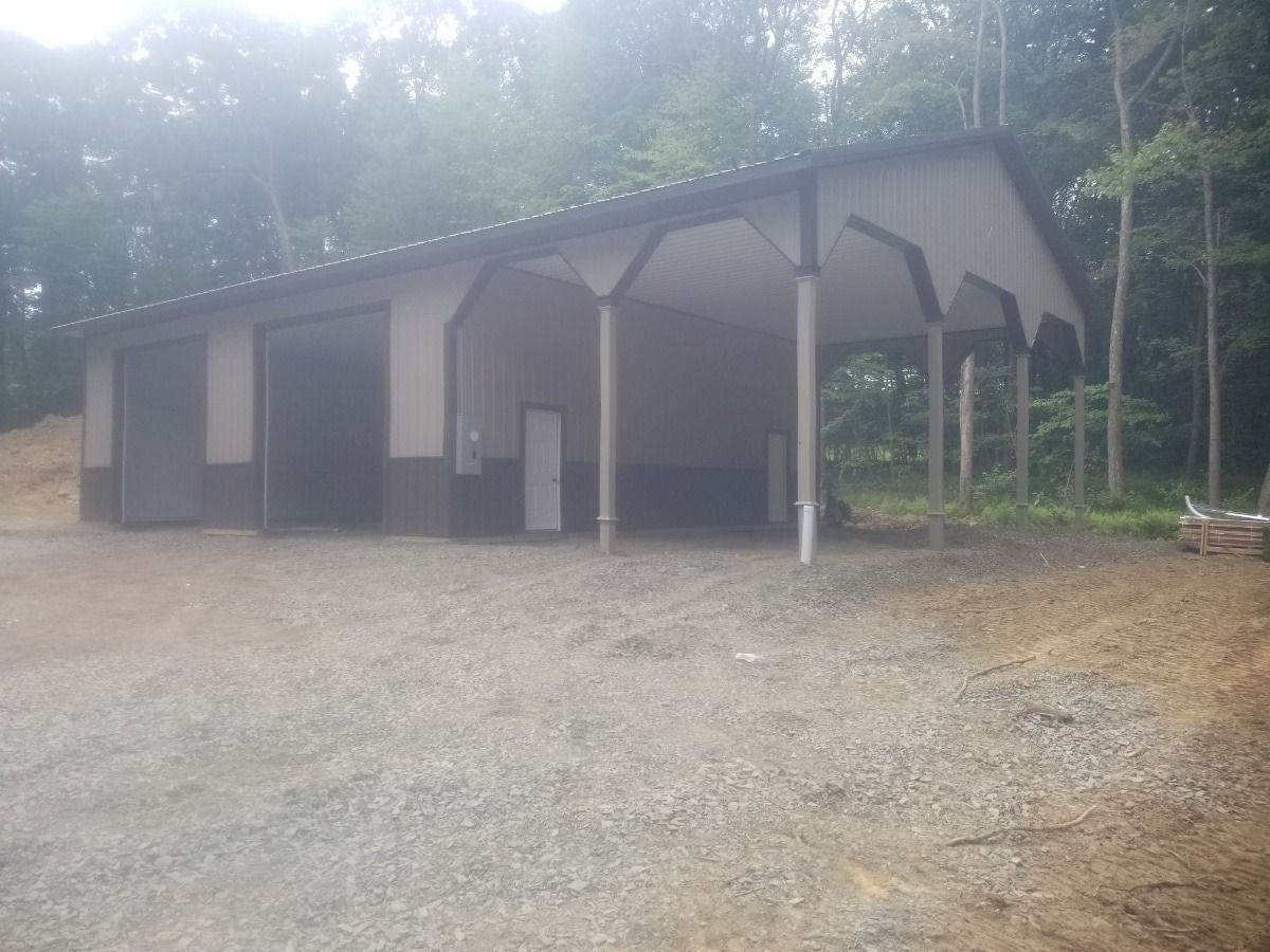 Gray metal shed with three garage bays on a gravel lot in a wooded area