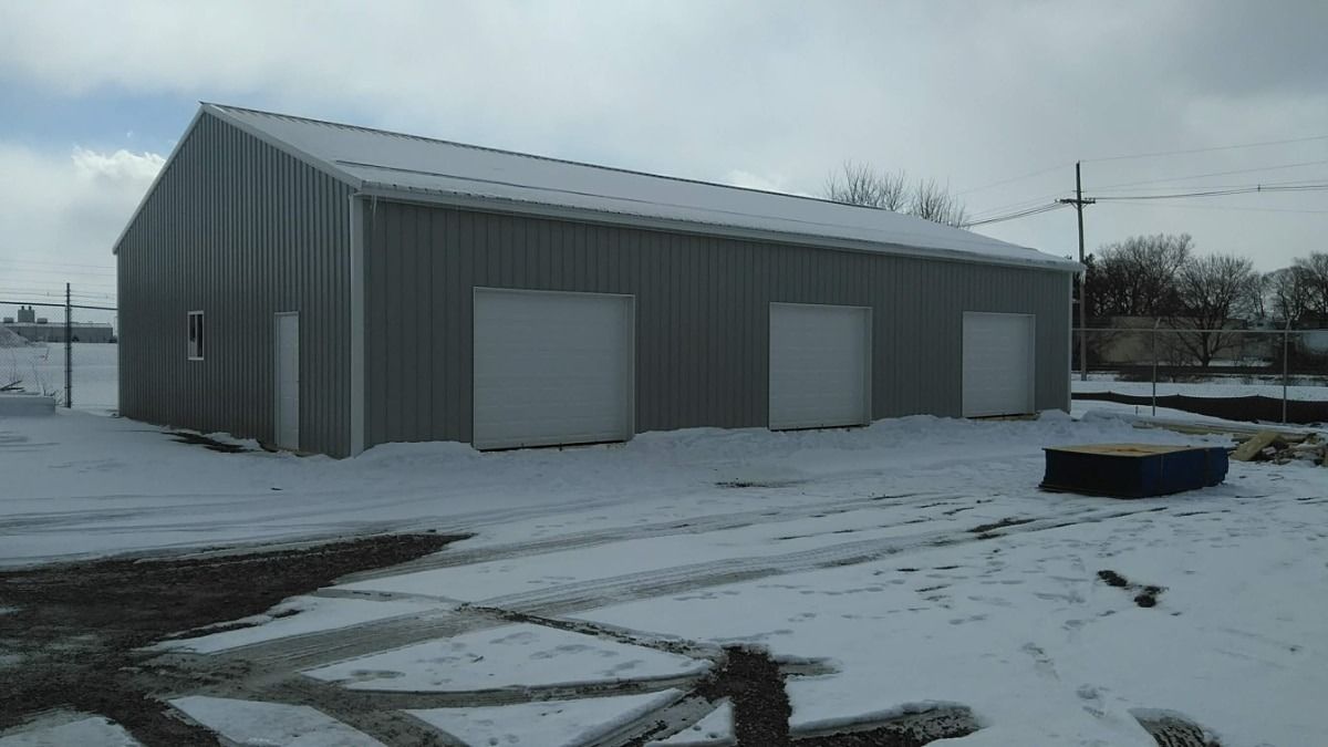 Snow-covered gray metal warehouse building with large garage doors on a cloudy day