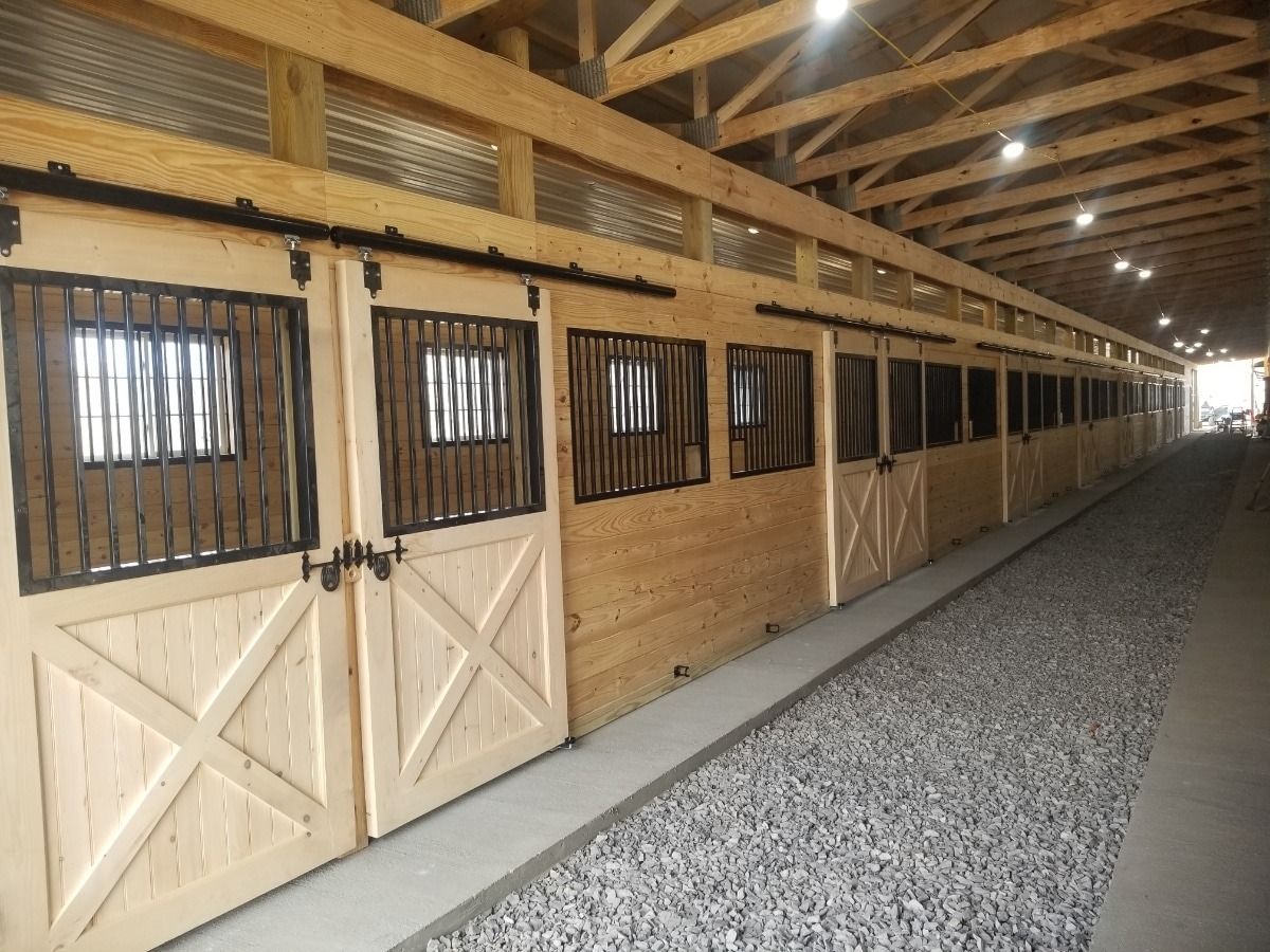 Wooden barn aisle with stalls on the left and a gravel floor under a vaulted roof.