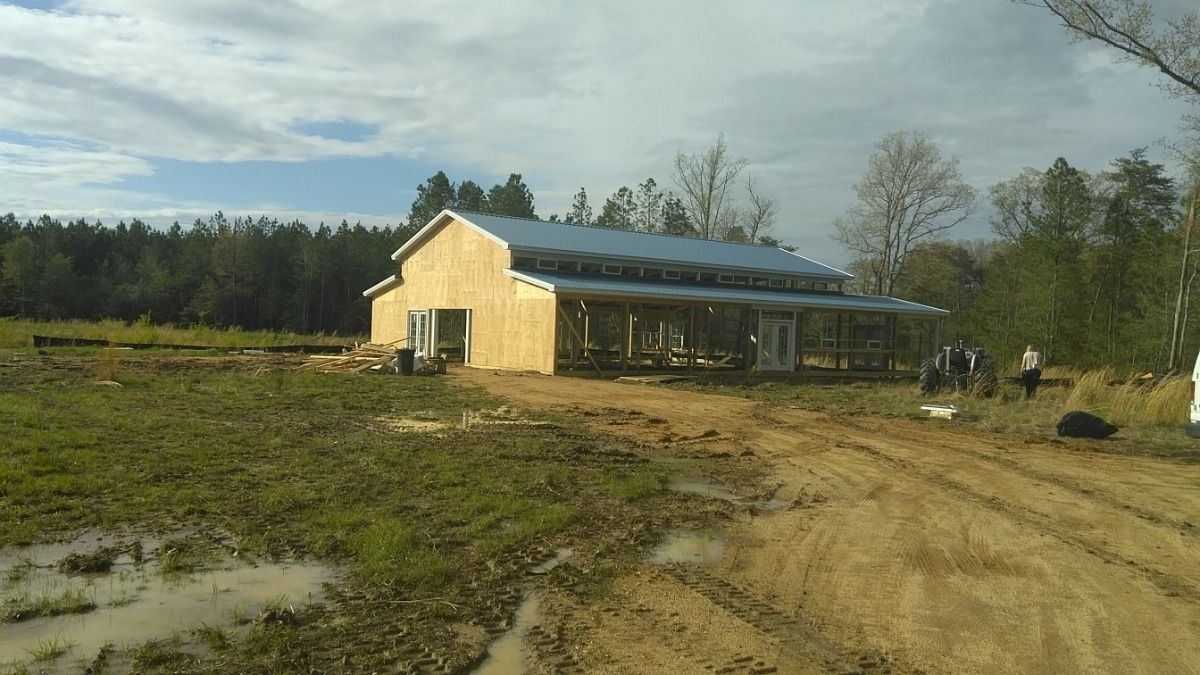 Rural farmhouse with a metal roof beside a dirt driveway and open field under a partly cloudy sky