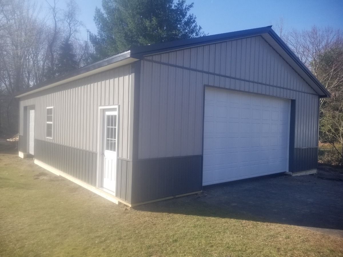 Gray metal garage/shed with white roll-up door and side entry, standing on a grassy lot in sunlight