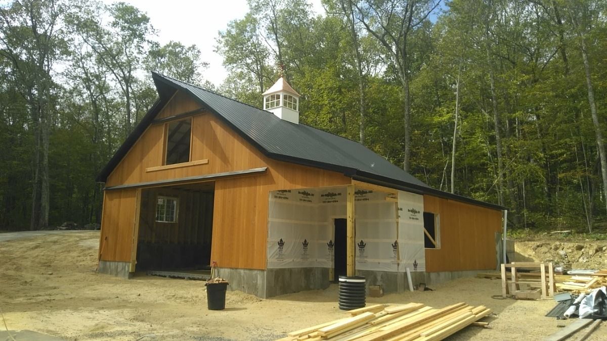 Wooden barn under construction in a wooded area, with building materials stacked outside.