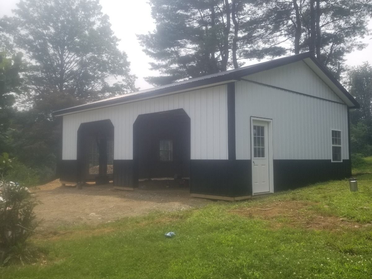 White metal barn with black trim and large garage doors on a grassy lot, surrounded by trees.