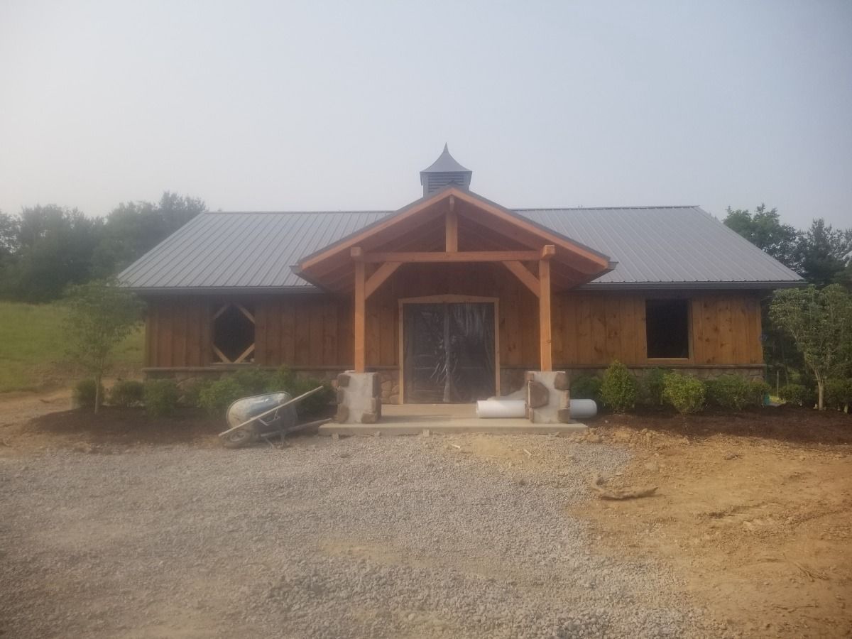 Rustic wooden cabin with a metal roof and central porch, set on a gravel drive in a rural landscape