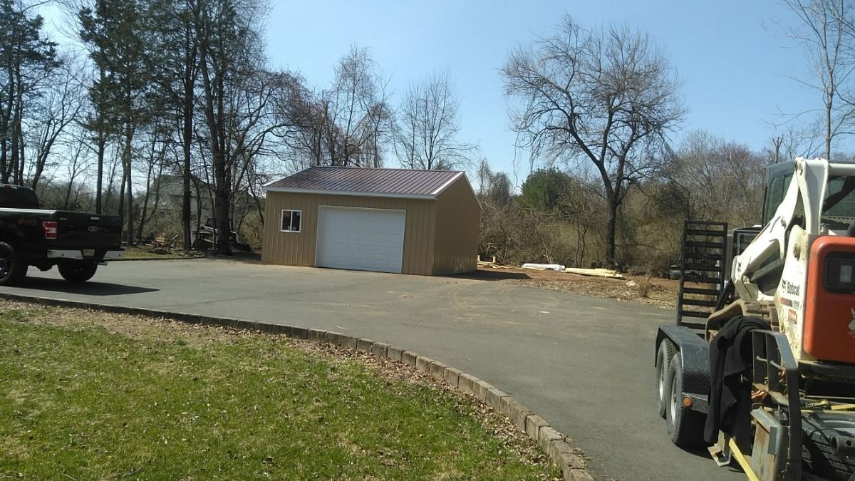 Driveway leading to a tan garage with a white door, trees around it, and a skid steer at the right edge.