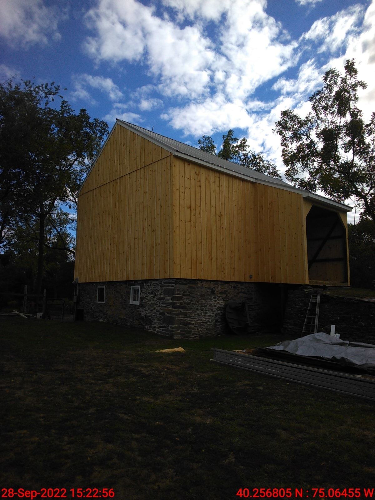 Yellow-sided barn with stone foundation under a cloudy blue sky, viewed from the yard.
