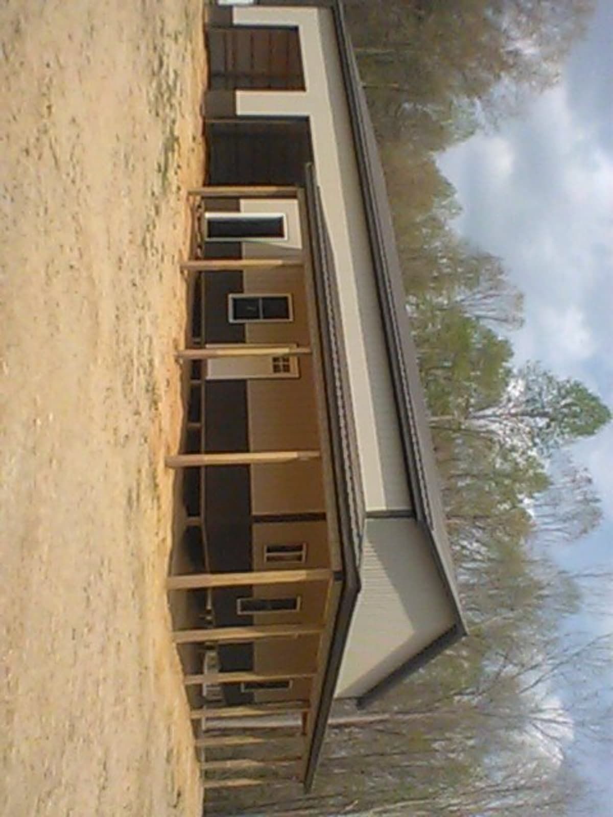 Single-story tan building with a covered porch and dark roof, seen from a dirt lot under a cloudy sky.