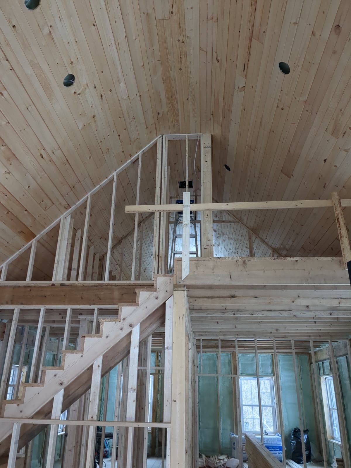 Interior of a wood-framed house under construction with stairs, loft railing, and vaulted pine ceiling